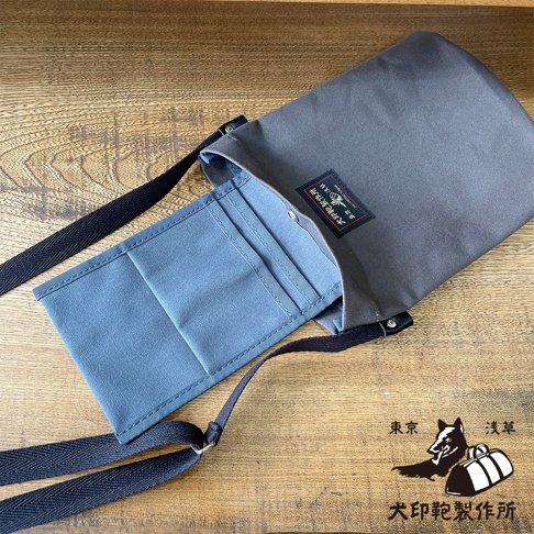 灰色帆布平板收納側背包，有兩個外部隔層，一個黑色肩帶，包上貼有印有日文的黑色標籤，右下角有狗和行李箱的標誌。