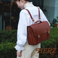 一名女子背著一個棕色皮革製成的三用公事包，包款設計極簡，配有兩個金屬扣環的翻蓋，側面有提把，肩帶可調整。