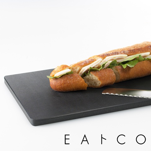 黑色合成材質砧板，表面有細微紋理，置於其上的是切成段的長棍麵包三明治，內夾雞肉、起司與生菜。一旁有一把鋸齒刀。