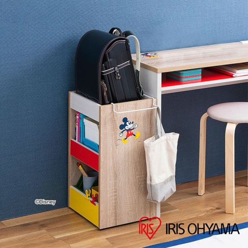 迪士尼系列木質收納櫃，木紋色，附滾輪，側面有米奇圖案，上方放置黑色書包，櫃內有書本、紅色和黃色抽屜，左下角有「©Disney」標誌。