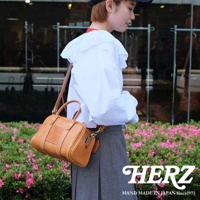 一位女性身著白色荷葉邊上衣和灰色裙子，斜挎一個焦糖色皮革迷你波士頓包，包上印有HERZ品牌標誌。