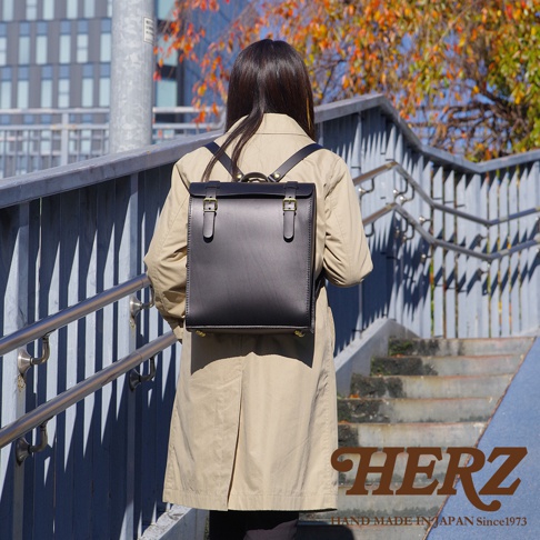 一位女性背著黑色厚實版皮革後背包，背包為方形設計，配有雙皮帶扣和金色金屬零件。