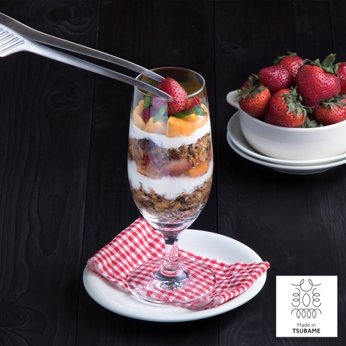 不鏽鋼材質的公筷夾，正在夾取杯中甜點裡的草莓，旁邊有裝滿草莓的白色碗。