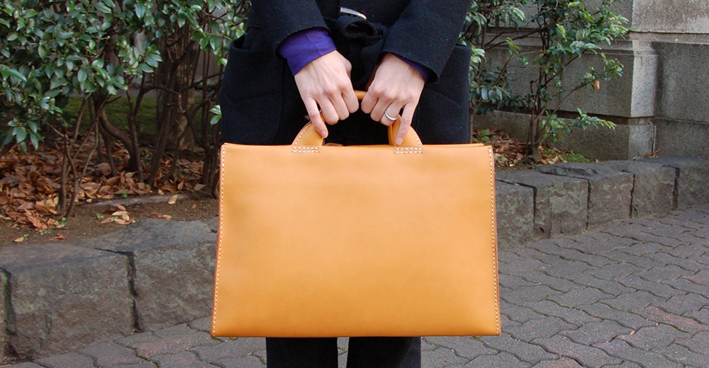 日本HERZ職人手作皮革簡約商務公事包，特寫一名女性雙手提著亮橘色光滑皮革公事包，背景為植被與石砌階梯。