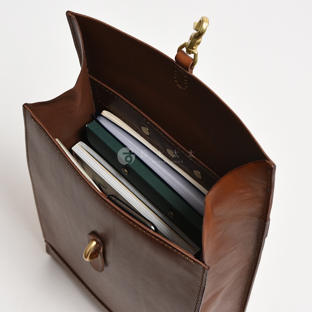 棕色皮革紙袋風後背包，內部裝有筆記本和文件，可見黃銅色金屬扣環和肩帶連接處。