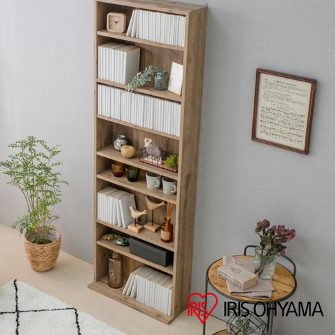 這是一款木紋質感的IRIS OHYAMA直立式開放書櫃，擺放於室內牆邊，層架上整齊排列著書籍、盆栽、馬克杯與裝飾品，牆面掛著一幅樂譜裝飾畫，右側設有圓形邊桌，整體營造出簡約溫馨的居家裝潢風格。