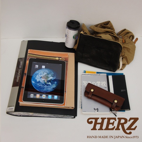 左側為黑色文件夾，上置一個裝有地球圖像的平板電腦，平板電腦被一個棕色皮革封套包覆，並有拉鍊設計。右側為一個黑色皮革手拿包，其後方有卡其色衣物，衣物旁放著一個印有星巴克標誌的白色環保杯。下方疊放著幾本筆記本，其中一本為淺灰色，書名為「寫真的學校」，其上放著一個棕色皮革筆夾。最下方為品牌標誌「HERZ」及「HAND MADE IN JAPAN Since1973」。