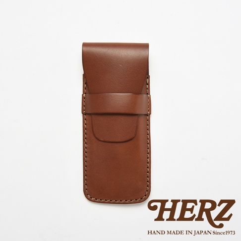 一款由日本品牌HERZ手工製作的棕色皮革鋼筆收納筆袋，採簡約的翻蓋式設計並以皮帶固定，表面呈現細緻的皮革紋理與手工縫線細節，下方印有品牌名稱與創立年份標誌。