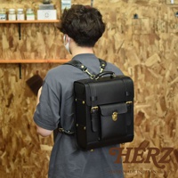 一位男士背著一個黑色皮革經典學院風後背包 R-100。背包為方形設計，採用全皮革材質，配有黃銅色金屬扣環、鎖扣及鉚釘裝飾。背包的正面有一個翻蓋小口袋，上方有提把，肩帶為寬版皮質設計。