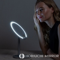 桌上型LED化妝鏡，圓形鏡面，邊緣環繞著明亮的LED燈光，鏡架與底座為銀灰色金屬材質，一位女士正在鏡前化妝。