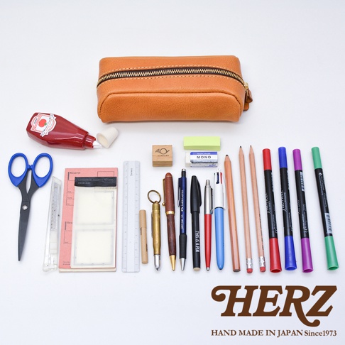 一個放在白色桌面上的日本HERZ職人手作棕色皮革收納筆袋，後方上方為筆袋本體，前方整齊排列著剪刀、裁紙刀、便條紙、尺、筆芯盒、各類書寫工具、鉛筆、彩色筆及橡皮擦，整體展現簡約優雅的文具收納風格。