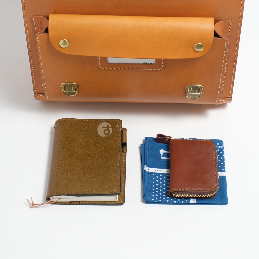 一個淺棕色的皮革橫式書包，下方擺放著一本棕色皮革筆記本和一個藍色圖案的布質小錢包。
