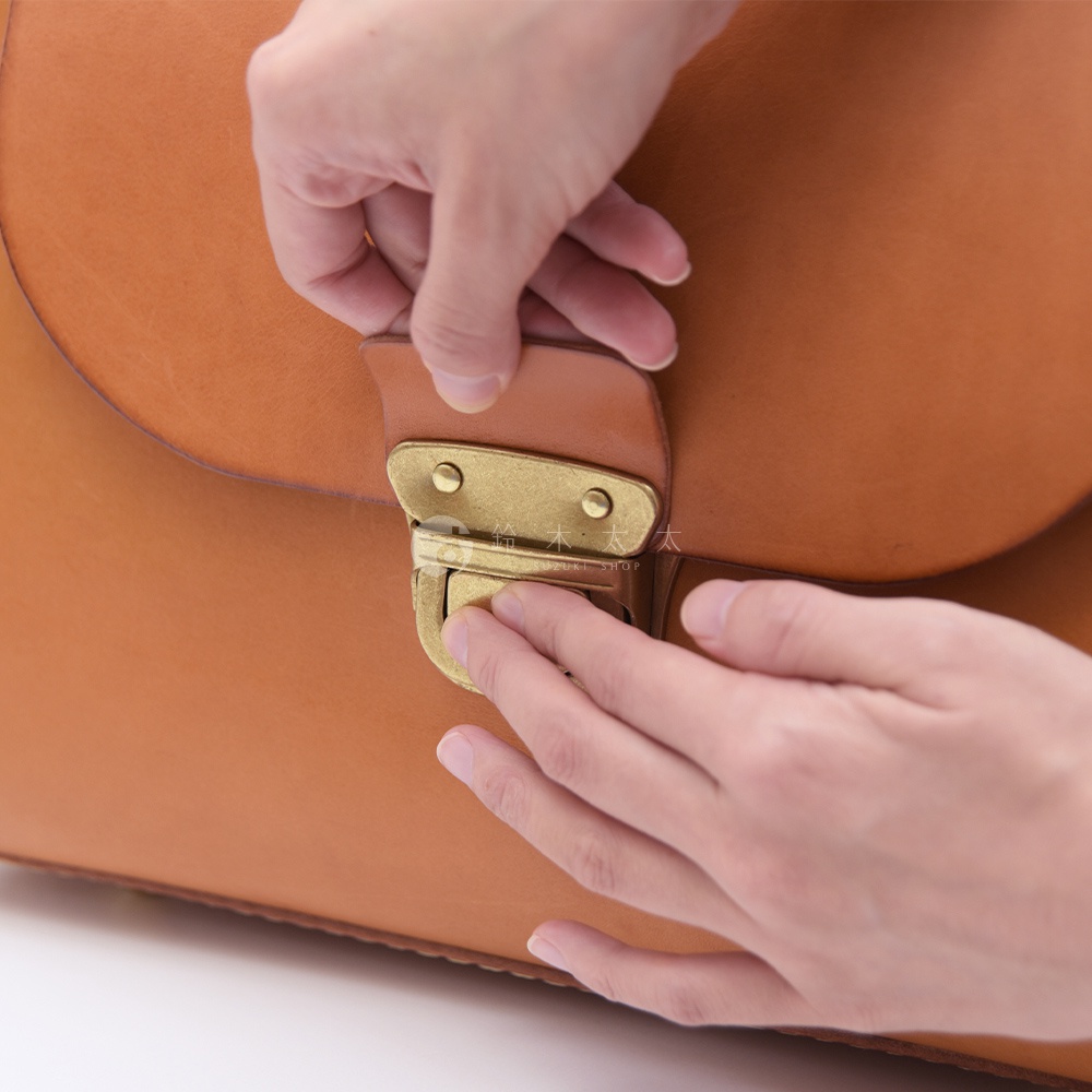 職人正在使用雙手調整一個復古黃銅鎖扣，鎖扣固定在一個由棕色皮革製成的包上，包上印有細小的日文與英文標誌，其中可見「SUZUKI SHOP」字樣。