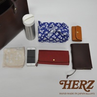 深棕色皮革直式托特包，旁邊擺放著不鏽鋼保溫瓶、印有貓咪圖案的藍色風呂敷、米白色繡花手帕、白色手機、紅色皮革長夾及深棕色皮革筆記本。