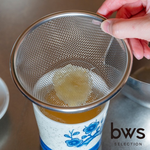 一隻手握著 bws SELECTION 不鏽鋼濾網，正將一塊蜂蜜放置於放置在帶有藍色花卉圖案的瓷杯上方的濾網中，濾網下方的杯子盛裝著金黃色的液體。