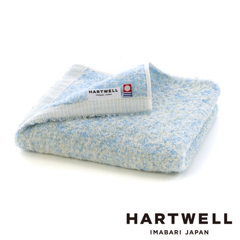 摺疊整齊的 Hartwell 今治毛巾，呈現淡雅的藍白混色質感，表面柔軟蓬鬆。毛巾上方設有標籤，印有 Hartwell Imabari Japan 字樣及專屬的今治毛巾認證標誌。整體色調清新柔和，展現高品質的編織細節與舒適親膚的視覺觸感。