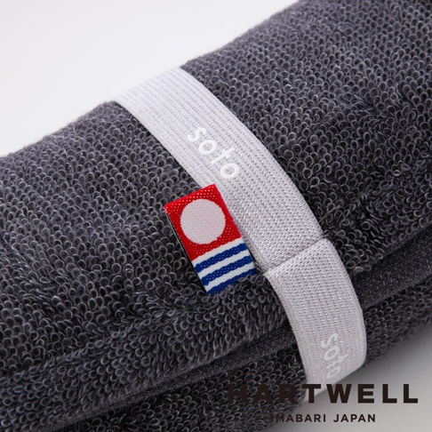 深灰色毛巾捲起，以白色束帶固定，束帶上印有 soto 字樣。毛巾旁有一個紅色標籤，標籤上有白色圓點和藍色條紋。