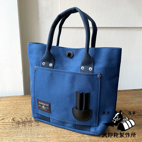 藍色帆布水桶托特包，設有深藍色織帶提把、外部貼袋和黑色皮革裝飾，袋身帶有品牌標籤。