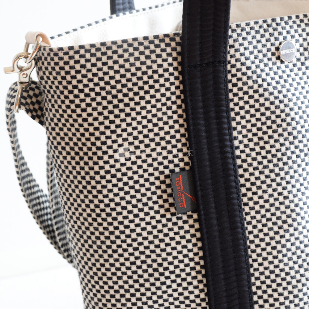 三河木綿刺子織兩用水桶包，織紋為黑白格紋，配有黑色皮革提把及可拆卸肩帶，包身上有紅色標籤。