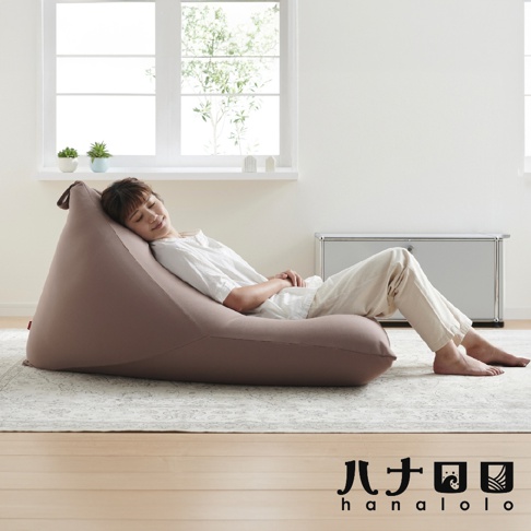 一名女子側臥於淺棕色針織布懶骨頭沙發上，沙發呈三角形，置於印花地毯上，背景為窗戶和金屬櫃。