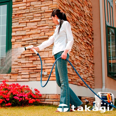 一位女士正使用白色噴槍和藍色水管為紅色花叢灑水，水管連接到一個帶有滾輪的白色捲軸。