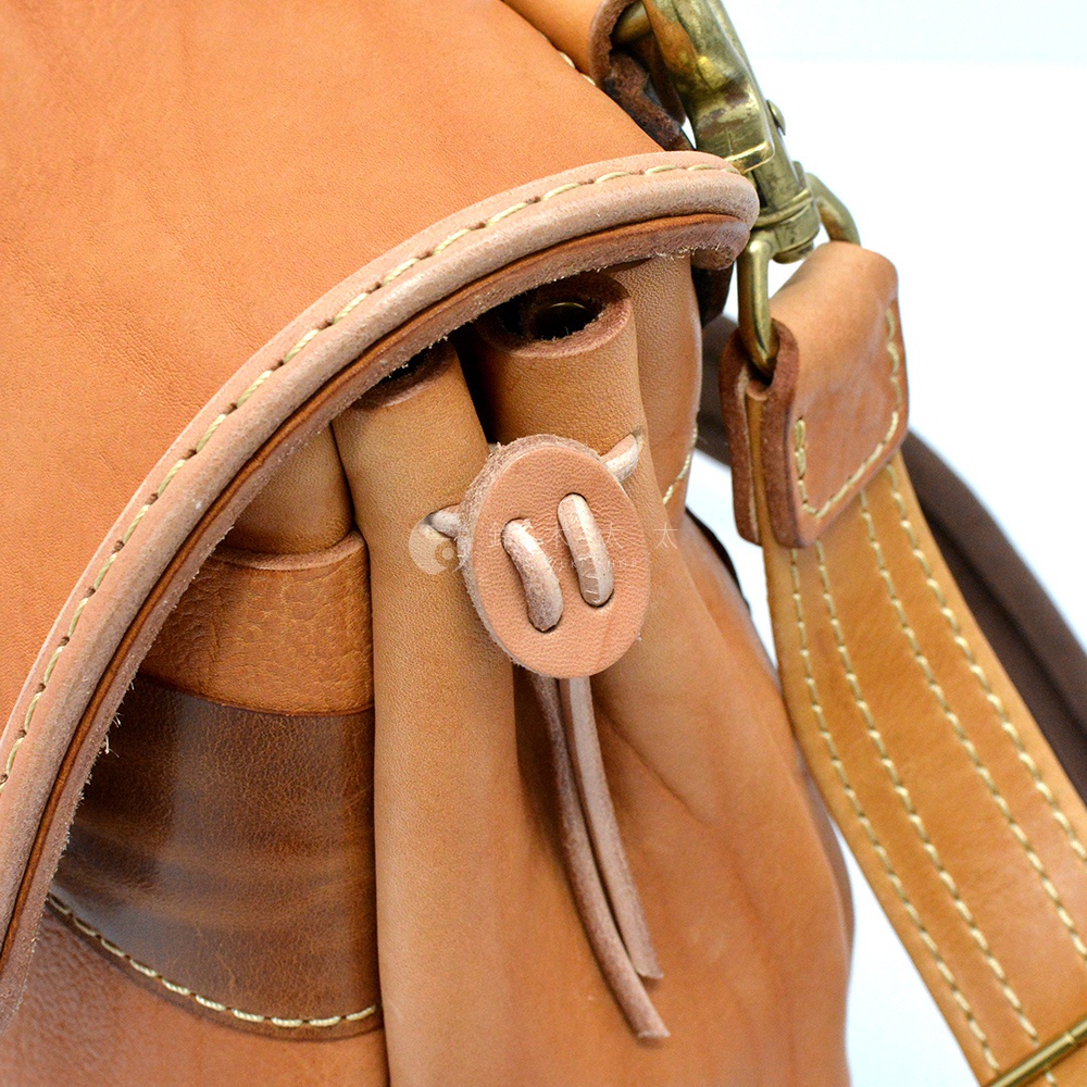 淺棕色皮革郵差包，特寫細節包含鹿角造型皮革束繩扣、車縫線、皮質提把及金屬掛鉤。