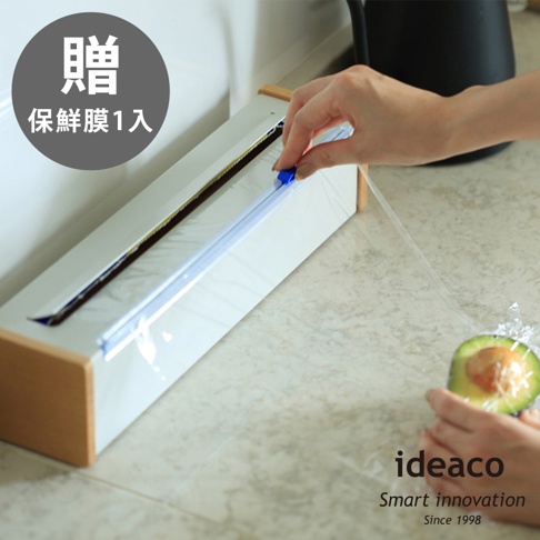 日本製原木鋼製保鮮膜切割器，白色主體搭配木質邊框，配有藍色切割滑軌，正在切割透明保鮮膜，左上角有「贈保鮮膜1入」的字樣。