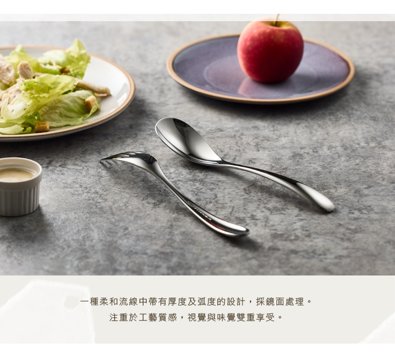 一組光滑的不鏽鋼湯匙和叉子，以鏡面處理，精緻的線條設計，擺放在石紋檯面上，一旁還有沙拉和一顆紅蘋果。