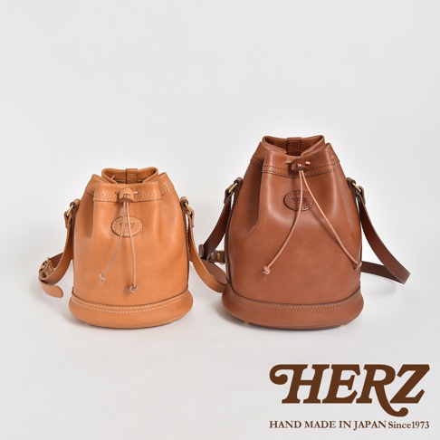 兩款職人手作皮革束口單肩包，左為淺棕色，右為深棕色。包身為圓筒狀，束口設計，附有皮革抽繩和可調整肩帶。材質應為牛皮，風格簡約。