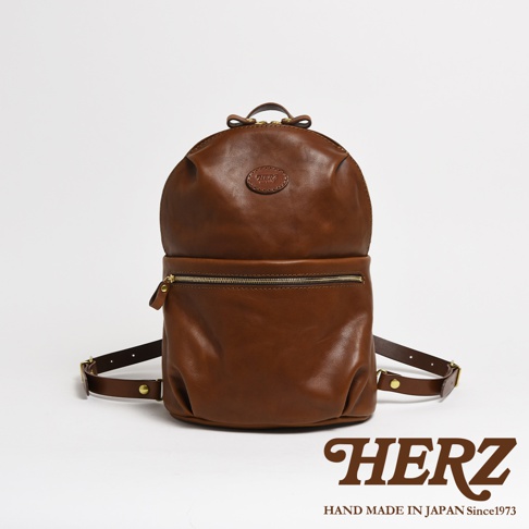 一棕色皮革後背包，圓潤的包身設計，正面有一個拉鍊口袋。包包上方有提把和肩帶，材質為光滑的皮革，配有金色拉鍊。