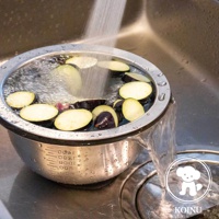 不鏽鋼濾水網與刻度料理盆，內有切片茄子，盆內裝有水，水流從濾水網上方傾瀉而下，盆外側有清晰的容量刻度標示。