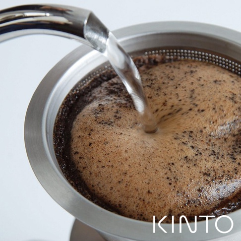 銀色不鏽鋼材質的免濾紙咖啡沖泡壺，壺口有細孔濾網，正有水流注入，咖啡粉與泡沫混合，呈現深棕色。