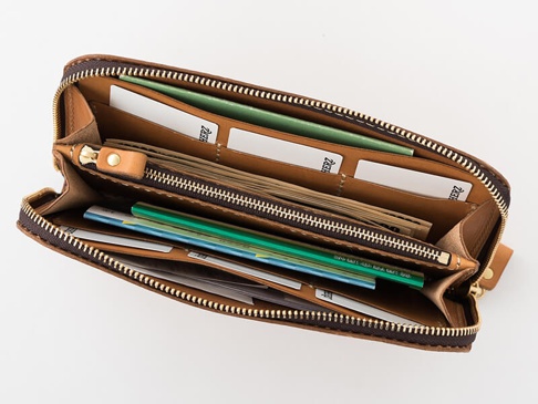 職人手作皮革大容量拉鍊長夾，呈現溫潤的淺棕色皮革，內部結構清晰，可見多個隔層、卡槽，並裝有紙鈔、卡片及綠色筆。拉鍊採用黃銅色金屬，質感良好。