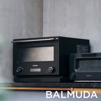 黑色 BALMUDA 微波烤箱，配有銀色提把、黑色玻璃門和數位顯示螢幕，左側有旋鈕和按鈕，右側另有一個黑色烤麵包機。