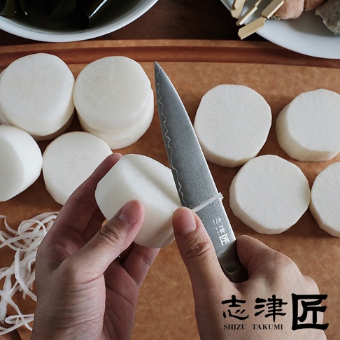 銀色不鏽鋼主廚刀，刀身有獨特紋路，正切著一片白色白蘿蔔，周圍散落幾片已切好的白蘿蔔圓片。