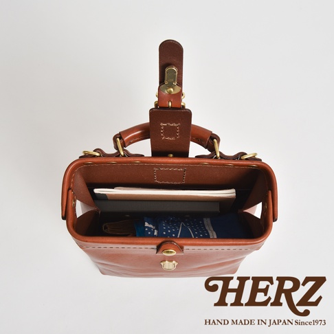 職人手作皮革迷你軟皮杜勒斯包，頂部視角，展示包內物品，含書本與藍色印花布料。包身為棕色皮革，配有金色金屬扣件與提把。