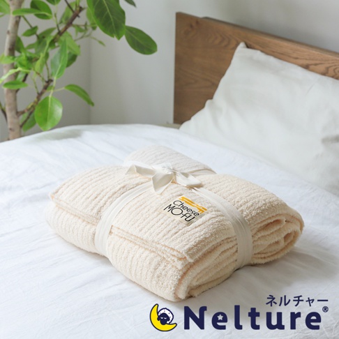 米白色絨毛毯，表面有細緻的條紋紋理，以白色緞帶繫綁，並附有標有「Cheese MOFU」的吊牌。毯子疊放在白色床單上，背景為木製床頭板和綠色植物。