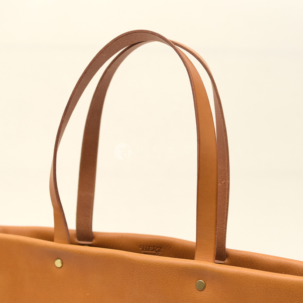 棕色皮革極簡托特包，有兩個對比色的提把，包身有兩個黃銅鉚釘，以及內側的「HERZ」品牌標誌。