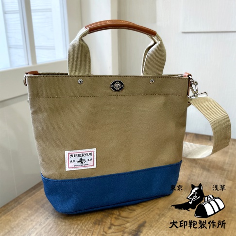 犬印純棉帆布兩用雙色托特包，上半部為卡其色，下半部為深藍色。包身側面有品牌標籤，提把及肩帶為米色，提把為皮革拼接設計，包包頂部有金屬釦和磁釦。
