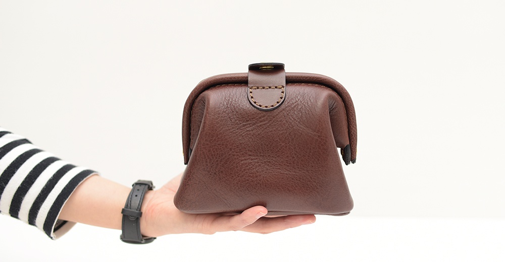 棕色皮革口金化妝包，有標誌性的縫線和金屬扣，由手持者展示。