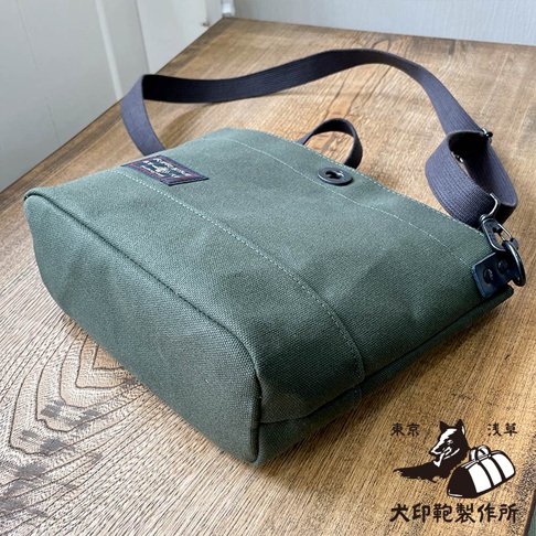 犬印鞄製作所的純綿帆布迷你側背包，採用日本防潑水帆布製作，深綠色包身，搭配深灰色的寬背帶，包蓋上有一個品牌標籤，右下角印有東京淺草犬印鞄製作所的標誌。