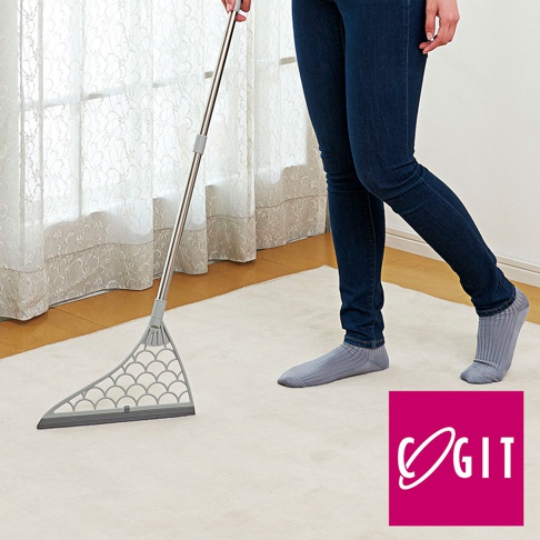 一隻手握著銀色金屬掃把，掃把頭為灰色，扇形設計，帶有圓形镂空圖案，底部有灰色膠條。掃帚置於淺米色地毯上，一位穿著深藍色牛仔褲和灰色條紋襪子的人站在右側。