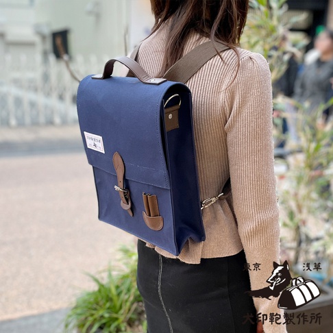 一款深藍色純棉帆布製成的直式三用側背包，配有棕色皮革飾邊、帶扣和提把。包包有兩條可調節的肩帶，其中一條斜挎在肩上，另一條則固定在身後。