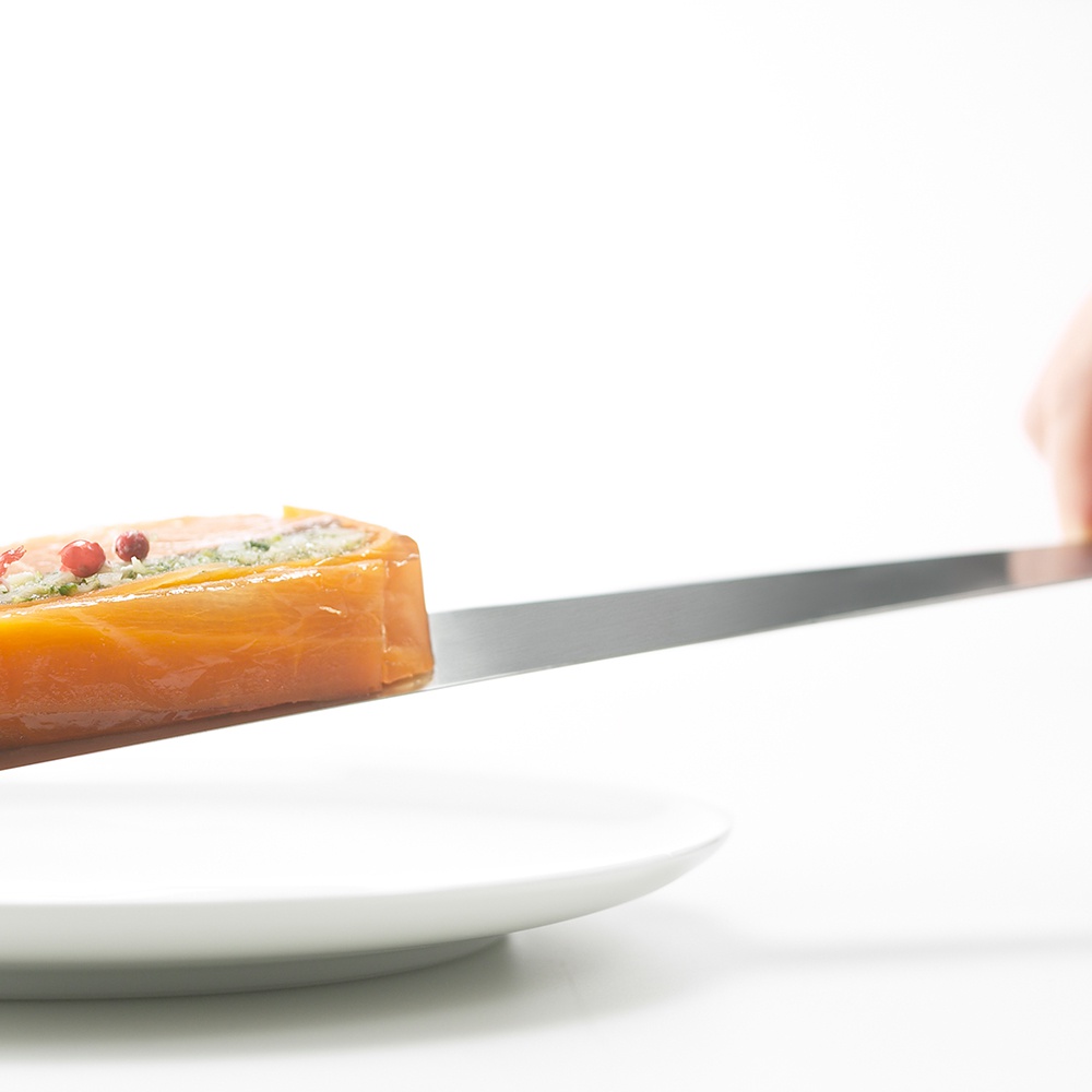 日製食物鏟，銀色金屬材質，輕巧薄片，搭配一塊帶有粉紅色胡椒粒和綠色香草的鮭魚料理，置於白色盤子上。