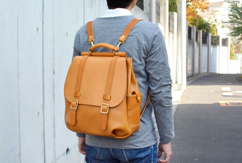 焦糖色皮革雙扣後背包，以瓦片狀的皮革覆蓋翻蓋設計，配有金屬扣環和可調節的皮革肩帶。