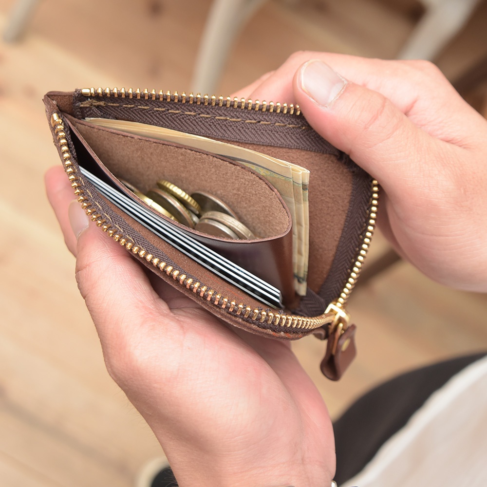 一雙手拿著一個棕色皮革L型拉鍊迷你短夾，夾內裝有信用卡、紙鈔和硬幣。