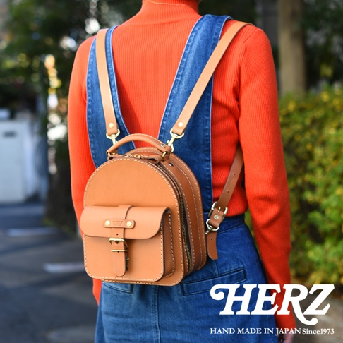 職人手作皮革迷你後背包，淺棕色，圓潤造型，主袋採用拉鍊開合，前方有一個帶有皮帶釦和金屬扣的翻蓋式口袋，整體為天然皮革材質，搭配可調式皮革肩帶。