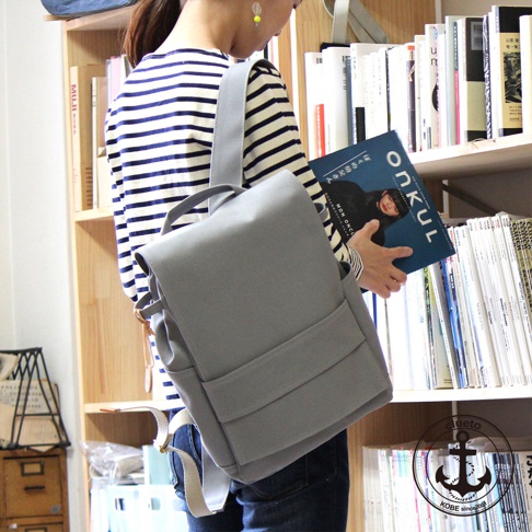 高島帆布前蓋式背包，灰色，配有可調節肩帶和外部口袋，一位身穿條紋上衣的女士肩背此包，身旁是擺滿書籍的書架。
