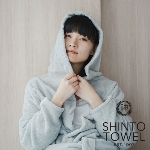 一位女性穿著淺藍色、蓬鬆柔軟的浴袍，浴袍帶有連帽設計，材質看起來是厚實的有機棉。