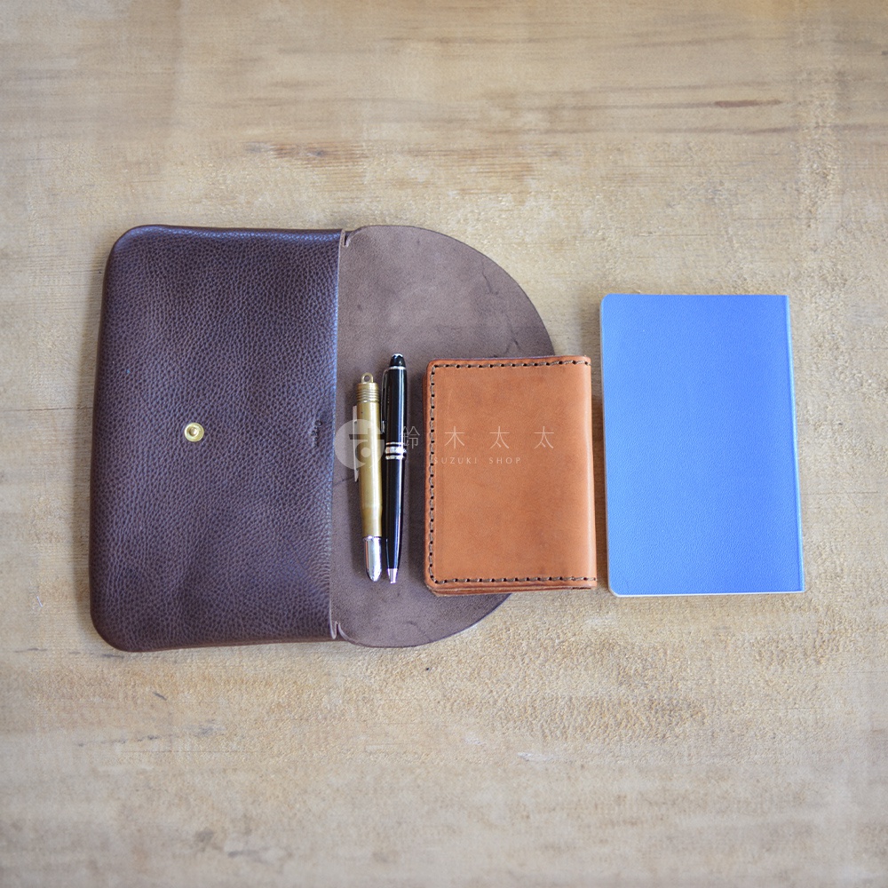 深棕色皮革製成的鈕扣式小物收納袋，內含一支金屬筆和一支黑色鋼筆，旁邊還放著一本橘棕色皮革封面筆記本和一本藍色筆記本。